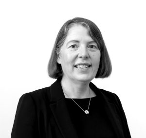 Black and white photo of Sarah Bushell. She is wearing smart black clothing and is smiling.
