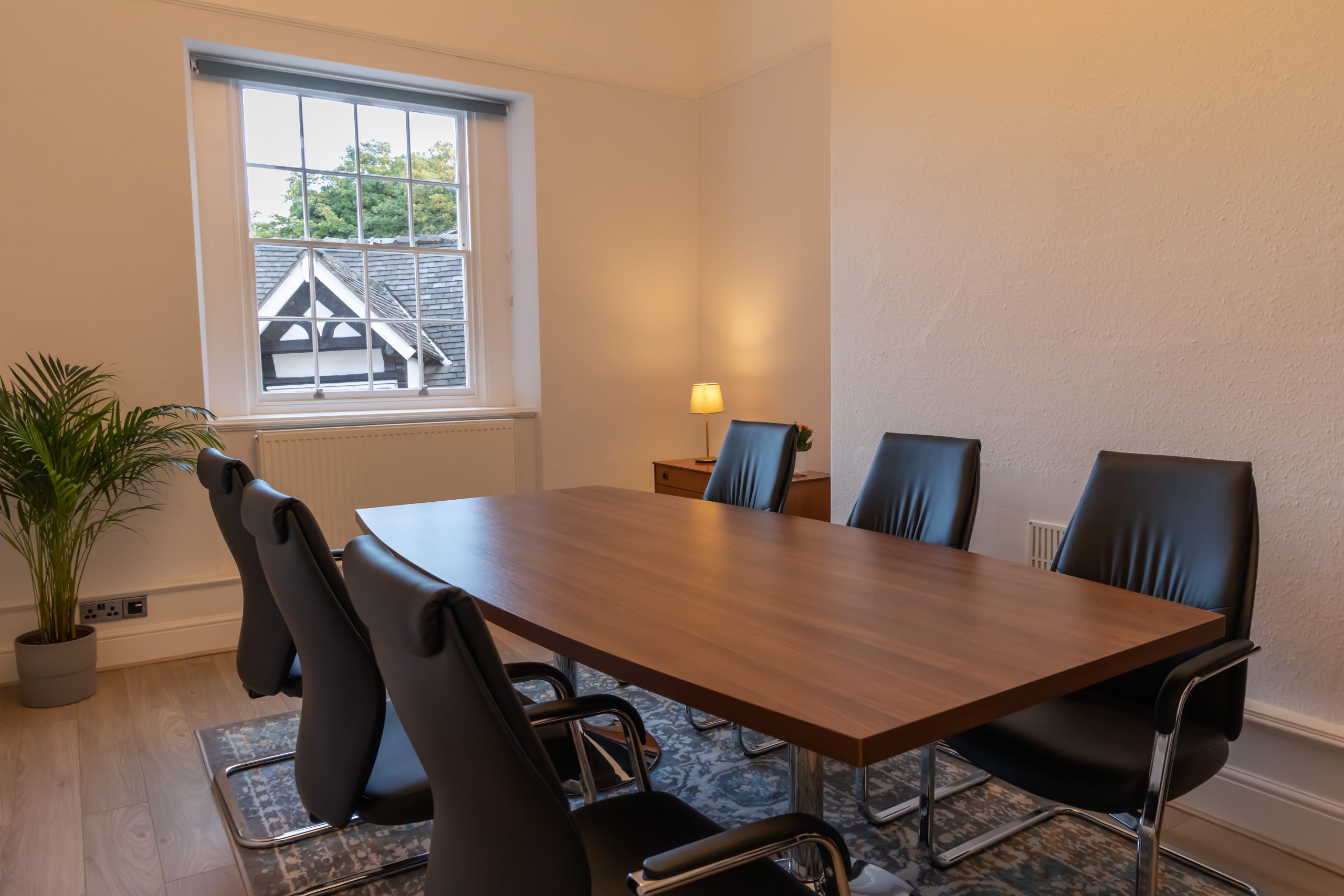 Photo of Spall Clark Solicitors office meeting room containing a wooden board room table and six black leather chairs. There is a blue and grey rug with orange and white accents on the light wooden floor with a green plant in the corner of the room. A small accent light is on. The room has high ceilings typical of a Georgian room. The sixteen panel window looks out onto Tudor rooftops of Little Street.
