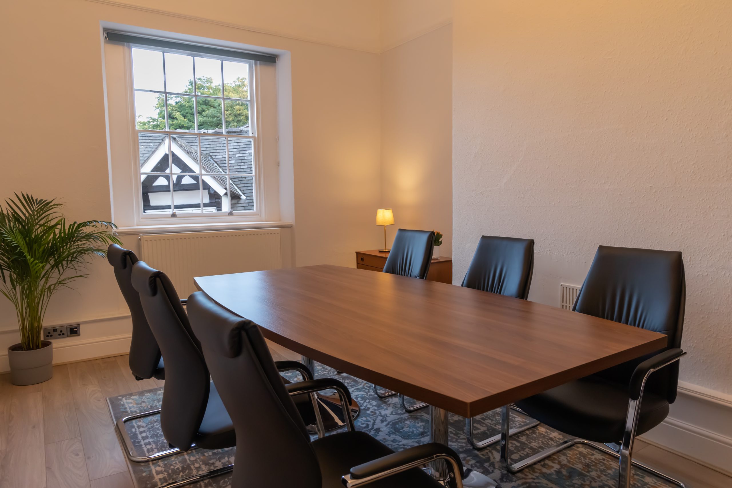 Photo of Spall Clark Solicitors office meeting room containing a wooden board room table and six black leather chairs. There is a blue and grey rug with orange and white accents on the light wooden floor with a green plant in the corner of the room. A small accent light is on. The room has high ceilings typical of a Georgian room. The sixteen panel window looks out onto Tudor rooftops of Little Street.