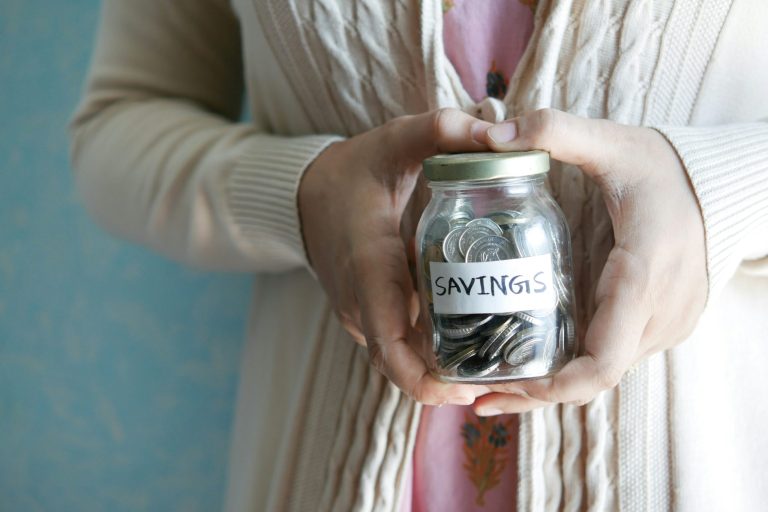 A jam jar of coins with a sticker labelled 'savings'. Held in two hands in a heart shape. Photo by Towfiqu Barbhuiya via Unsplash.com