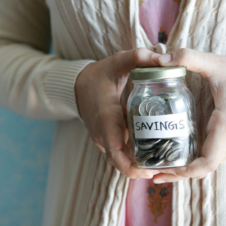 A jam jar of coins with a sticker labelled 'savings'. Held in two hands in a heart shape. Photo by Towfiqu Barbhuiya via Unsplash.com