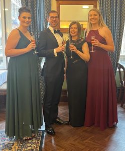 Melissa Howman (green dress), Paul Clark (dinner jacket), Becky Spall (black dress) and Natasha Yelland (red dress) in evening wear at the St James Club, Standing in front of a window with champagne.