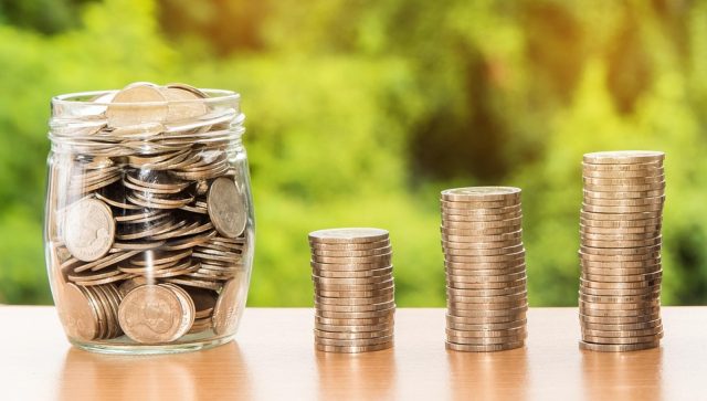 Money Coins in a jar with three small stacks of coins next to the jar. Each stack is slightly larger than the previous.